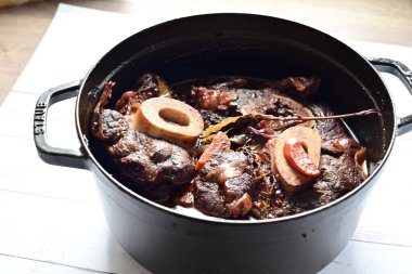 Kırmızı şarapta pişirilmiş kemikli yumuşak sığır eti, taze otlar ve tek bir kırsal siyah tencerede kök sebzelerle servis edilir..