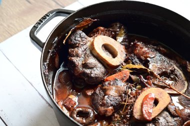 Kırmızı şarapta pişirilmiş kemikli yumuşak sığır eti, taze otlar ve tek bir kırsal siyah tencerede kök sebzelerle servis edilir..