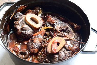 Kırmızı şarapta pişirilmiş kemikli yumuşak sığır eti, taze otlar ve tek bir kırsal siyah tencerede kök sebzelerle servis edilir..