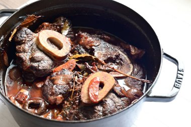 Kırmızı şarapta pişirilmiş kemikli yumuşak sığır eti, taze otlar ve tek bir kırsal siyah tencerede kök sebzelerle servis edilir..