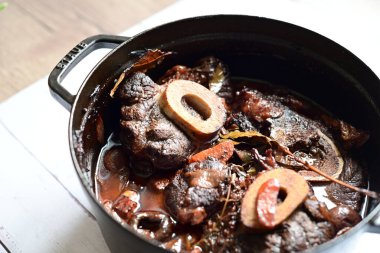 Kırmızı şarapta pişirilmiş kemikli yumuşak sığır eti, taze otlar ve tek bir kırsal siyah tencerede kök sebzelerle servis edilir..