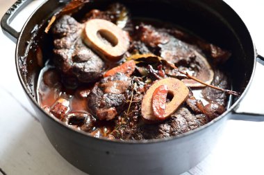 Kırmızı şarapta pişirilmiş kemikli yumuşak sığır eti, taze otlar ve tek bir kırsal siyah tencerede kök sebzelerle servis edilir..