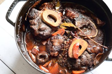 Kırmızı şarapta pişirilmiş kemikli yumuşak sığır eti, taze otlar ve tek bir kırsal siyah tencerede kök sebzelerle servis edilir..