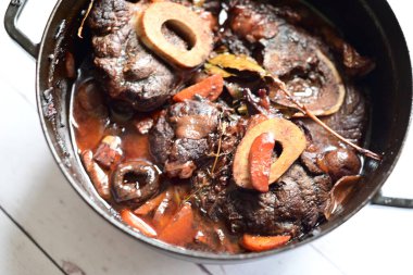 Kırmızı şarapta pişirilmiş kemikli yumuşak sığır eti, taze otlar ve tek bir kırsal siyah tencerede kök sebzelerle servis edilir..