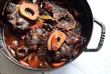 Kırmızı şarapta pişirilmiş kemikli yumuşak sığır eti, taze otlar ve tek bir kırsal siyah tencerede kök sebzelerle servis edilir..