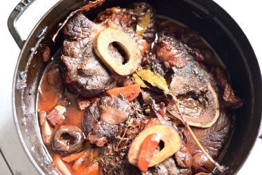 Kırmızı şarapta pişirilmiş kemikli yumuşak sığır eti, taze otlar ve tek bir kırsal siyah tencerede kök sebzelerle servis edilir..