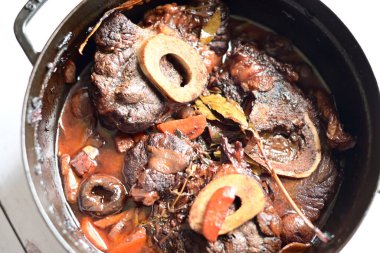 Kırmızı şarapta pişirilmiş kemikli yumuşak sığır eti, taze otlar ve tek bir kırsal siyah tencerede kök sebzelerle servis edilir..