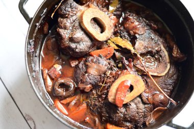 Kırmızı şarapta pişirilmiş kemikli yumuşak sığır eti, taze otlar ve tek bir kırsal siyah tencerede kök sebzelerle servis edilir..