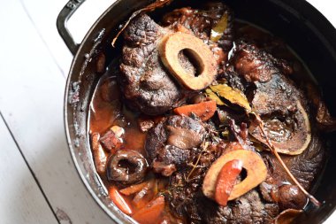 Kırmızı şarapta pişirilmiş kemikli yumuşak sığır eti, taze otlar ve tek bir kırsal siyah tencerede kök sebzelerle servis edilir..