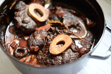 Kırmızı şarapla pişirilmiş kemikli yumuşak sığır eti, taze otlar ve tek bir kırsal siyah tencerede kök sebzelerle servis edilir. Zengin sos ve yavaş pişirilmiş malzemeler geleneksel bir konforlu yemek yemeğinin altını çiziyor..