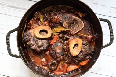 Kırmızı şarapta pişirilmiş kemikli yumuşak sığır eti, taze otlar ve tek bir kırsal siyah tencerede kök sebzelerle servis edilir..