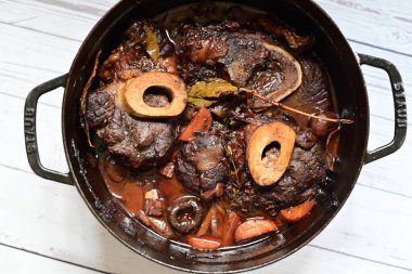 Kırmızı şarapta pişirilmiş kemikli yumuşak sığır eti, taze otlar ve tek bir kırsal siyah tencerede kök sebzelerle servis edilir..