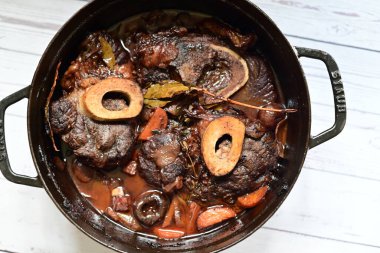 Kırmızı şarapta pişirilmiş kemikli yumuşak sığır eti, taze otlar ve tek bir kırsal siyah tencerede kök sebzelerle servis edilir..