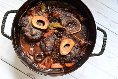 Kırmızı şarapta pişirilmiş kemikli yumuşak sığır eti, taze otlar ve tek bir kırsal siyah tencerede kök sebzelerle servis edilir..