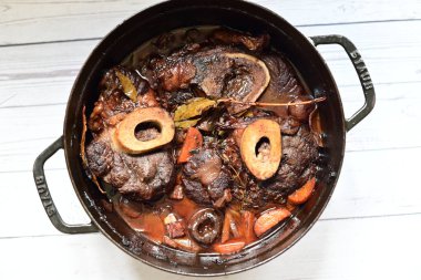 Kırmızı şarapta pişirilmiş kemikli yumuşak sığır eti, taze otlar ve tek bir kırsal siyah tencerede kök sebzelerle servis edilir..