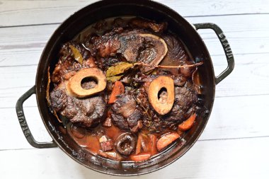 Kırmızı şarapta pişirilmiş kemikli yumuşak sığır eti, taze otlar ve tek bir kırsal siyah tencerede kök sebzelerle servis edilir..