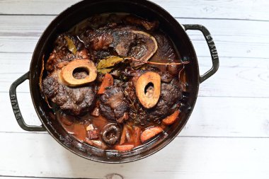 Kırmızı şarapta pişirilmiş kemikli yumuşak sığır eti, taze otlar ve tek bir kırsal siyah tencerede kök sebzelerle servis edilir..