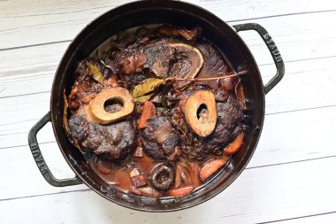 Kırmızı şarapta pişirilmiş kemikli yumuşak sığır eti, taze otlar ve tek bir kırsal siyah tencerede kök sebzelerle servis edilir..