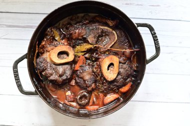 Kırmızı şarapta pişirilmiş kemikli yumuşak sığır eti, taze otlar ve tek bir kırsal siyah tencerede kök sebzelerle servis edilir..