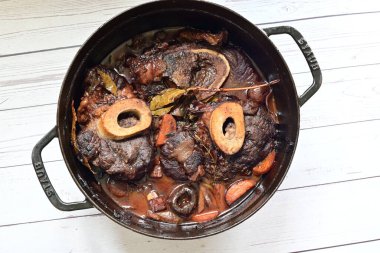 Kırmızı şarapta pişirilmiş kemikli yumuşak sığır eti, taze otlar ve tek bir kırsal siyah tencerede kök sebzelerle servis edilir..