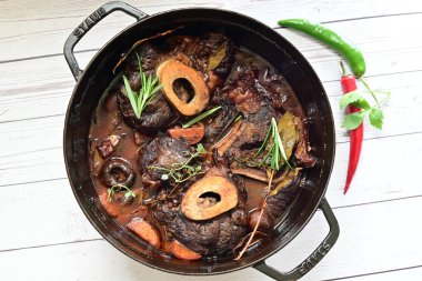 Kırmızı şarapta pişirilmiş kemikli yumuşak sığır eti, taze otlar ve tek bir kırsal siyah tencerede kök sebzelerle servis edilir..