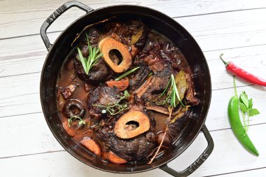 Kırmızı şarapta pişirilmiş kemikli yumuşak sığır eti, taze otlar ve tek bir kırsal siyah tencerede kök sebzelerle servis edilir..