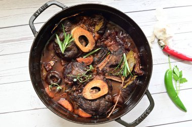 Kırmızı şarapta pişirilmiş kemikli yumuşak sığır eti, taze otlar ve tek bir kırsal siyah tencerede kök sebzelerle servis edilir..