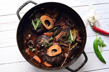 Kırmızı şarapta pişirilmiş kemikli yumuşak sığır eti, taze otlar ve tek bir kırsal siyah tencerede kök sebzelerle servis edilir..