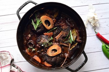 Kırmızı şarapta pişirilmiş kemikli yumuşak sığır eti, taze otlar ve tek bir kırsal siyah tencerede kök sebzelerle servis edilir..