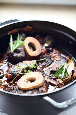 Kırmızı şarapta pişirilmiş kemikli yumuşak sığır eti, taze otlar ve tek bir kırsal siyah tencerede kök sebzelerle servis edilir..