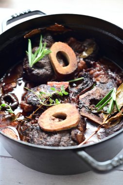 Kırmızı şarapta pişirilmiş kemikli yumuşak sığır eti, taze otlar ve tek bir kırsal siyah tencerede kök sebzelerle servis edilir..