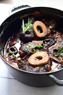 Kırmızı şarapta pişirilmiş kemikli yumuşak sığır eti, taze otlar ve tek bir kırsal siyah tencerede kök sebzelerle servis edilir..