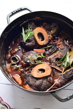 Kırmızı şarapta pişirilmiş kemikli yumuşak sığır eti, taze otlar ve tek bir kırsal siyah tencerede kök sebzelerle servis edilir..