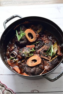 Yanında taze patates, kök sebze ve taze otlarla birlikte kırmızı şarap soslu kemikli haşlanmış sığır eti servis edilir. Beyaz bir tabakta parlak bir arkaplanda sunulan bu çanak, klasik konforlu yiyecekleri inceliyor.