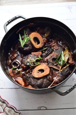 Yanında taze patates, kök sebze ve taze otlarla birlikte kırmızı şarap soslu kemikli haşlanmış sığır eti servis edilir. Beyaz bir tabakta parlak bir arkaplanda sunulan bu çanak, klasik konforlu yiyecekleri inceliyor.