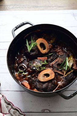 Yanında taze patates, kök sebze ve taze otlarla birlikte kırmızı şarap soslu kemikli haşlanmış sığır eti servis edilir. Beyaz bir tabakta parlak bir arkaplanda sunulan bu çanak, klasik konforlu yiyecekleri inceliyor.