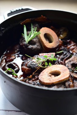 Yanında taze patates, kök sebze ve taze otlarla birlikte kırmızı şarap soslu kemikli haşlanmış sığır eti servis edilir. Beyaz bir tabakta parlak bir arkaplanda sunulan bu çanak, klasik konforlu yiyecekleri inceliyor.