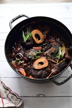 Yanında taze patates, kök sebze ve taze otlarla birlikte kırmızı şarap soslu kemikli haşlanmış sığır eti servis edilir. Beyaz bir tabakta parlak bir arkaplanda sunulan bu çanak, klasik konforlu yiyecekleri inceliyor.