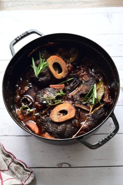 Yanında taze patates, kök sebze ve taze otlarla birlikte kırmızı şarap soslu kemikli haşlanmış sığır eti servis edilir. Beyaz bir tabakta parlak bir arkaplanda sunulan bu çanak, klasik konforlu yiyecekleri inceliyor.