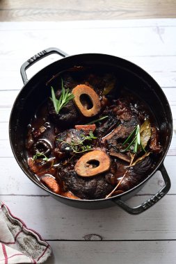 Yanında taze patates, kök sebze ve taze otlarla birlikte kırmızı şarap soslu kemikli haşlanmış sığır eti servis edilir. Beyaz bir tabakta parlak bir arkaplanda sunulan bu çanak, klasik konforlu yiyecekleri inceliyor.