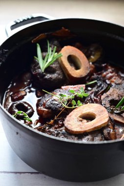 Yanında taze patates, kök sebze ve taze otlarla birlikte kırmızı şarap soslu kemikli haşlanmış sığır eti servis edilir. Beyaz bir tabakta parlak bir arkaplanda sunulan bu çanak, klasik konforlu yiyecekleri inceliyor.