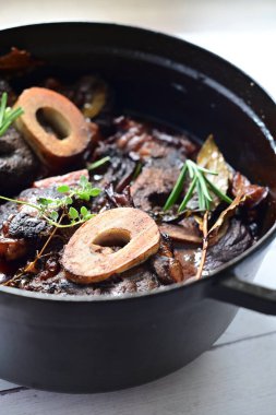 Yanında taze patates, kök sebze ve taze otlarla birlikte kırmızı şarap soslu kemikli haşlanmış sığır eti servis edilir. Beyaz bir tabakta parlak bir arkaplanda sunulan bu çanak, klasik konforlu yiyecekleri inceliyor.