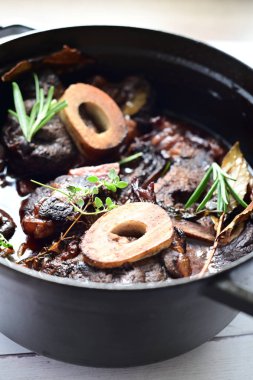 Yanında taze patates, kök sebze ve taze otlarla birlikte kırmızı şarap soslu kemikli haşlanmış sığır eti servis edilir. Beyaz bir tabakta parlak bir arkaplanda sunulan bu çanak, klasik konforlu yiyecekleri inceliyor.