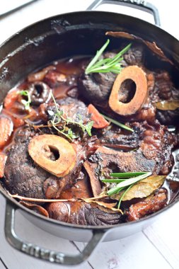 Yanında taze patates, kök sebze ve taze otlarla birlikte kırmızı şarap soslu kemikli haşlanmış sığır eti servis edilir. Beyaz bir tabakta parlak bir arkaplanda sunulan bu çanak, klasik konforlu yiyecekleri inceliyor.