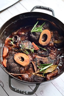 Yanında taze patates, kök sebze ve taze otlarla birlikte kırmızı şarap soslu kemikli haşlanmış sığır eti servis edilir. Beyaz bir tabakta parlak bir arkaplanda sunulan bu çanak, klasik konforlu yiyecekleri inceliyor.