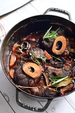 Yanında taze patates, kök sebze ve taze otlarla birlikte kırmızı şarap soslu kemikli haşlanmış sığır eti servis edilir. Beyaz bir tabakta parlak bir arkaplanda sunulan bu çanak, klasik konforlu yiyecekleri inceliyor.