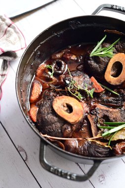 Yanında taze patates, kök sebze ve taze otlarla birlikte kırmızı şarap soslu kemikli haşlanmış sığır eti servis edilir. Beyaz bir tabakta parlak bir arkaplanda sunulan bu çanak, klasik konforlu yiyecekleri inceliyor.