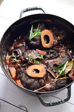 Yanında taze patates, kök sebze ve taze otlarla birlikte kırmızı şarap soslu kemikli haşlanmış sığır eti servis edilir. Beyaz bir tabakta parlak bir arkaplanda sunulan bu çanak, klasik konforlu yiyecekleri inceliyor.