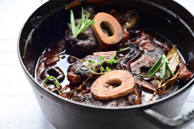 Yanında taze patates, kök sebze ve taze otlarla birlikte kırmızı şarap soslu kemikli haşlanmış sığır eti servis edilir. Beyaz bir tabakta parlak bir arkaplanda sunulan bu çanak, klasik konforlu yiyecekleri inceliyor.