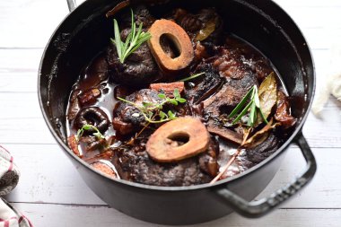 Yanında taze patates, kök sebze ve taze otlarla birlikte kırmızı şarap soslu kemikli haşlanmış sığır eti servis edilir. Beyaz bir tabakta parlak bir arkaplanda sunulan bu çanak, klasik konforlu yiyecekleri inceliyor.